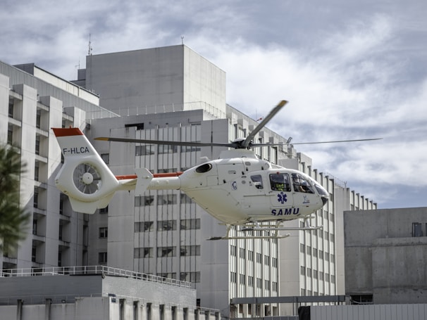 A medical helicopter is in mid-flight near a large, modern hospital building. The helicopter is white with red accents and displays the word 'SAMU' on its side, indicating it is an emergency medical service vehicle. The background consists of the hospital's geometric, rectangular architecture under a cloudy sky.