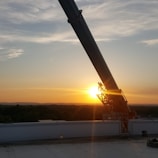 A large telescope or observatory structure is silhouetted against a vibrant sunset. The sky is painted with hues of orange, pink, and blue, while the sun is positioned near the horizon, casting a warm glow. In the foreground, a flat rooftop or platform is partially visible.