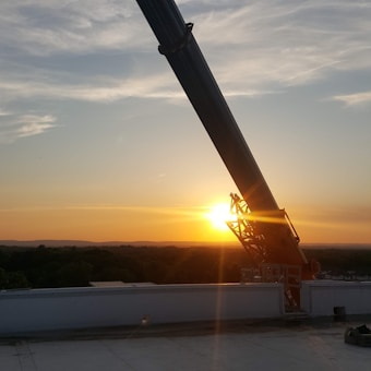 A large telescope or observatory structure is silhouetted against a vibrant sunset. The sky is painted with hues of orange, pink, and blue, while the sun is positioned near the horizon, casting a warm glow. In the foreground, a flat rooftop or platform is partially visible.