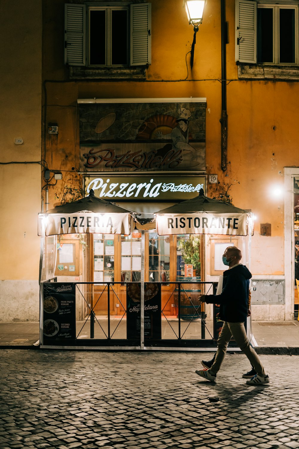 Uomo in giacca nera e pantaloni neri in piedi davanti al negozio di legno marrone durante il giorno