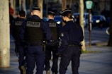 Several uniformed police officers walking along a city street. They are wearing dark uniforms and protective vests, with one carrying a communication device. Urban setting with parked cars and street signs in the background.