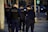 Several uniformed police officers walking along a city street. They are wearing dark uniforms and protective vests, with one carrying a communication device. Urban setting with parked cars and street signs in the background.