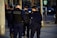 Several uniformed police officers walking along a city street. They are wearing dark uniforms and protective vests, with one carrying a communication device. Urban setting with parked cars and street signs in the background.