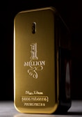 A gold-colored, rectangular perfume bottle with rounded edges and embossed text on its surface. The text includes '1 Million' along with the brand name and volume details. The bottle is set against a dark background, highlighting its shiny metallic finish.