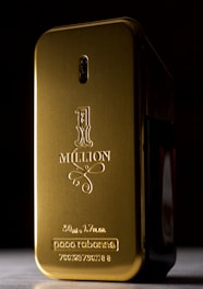 A gold-colored, rectangular perfume bottle with rounded edges and embossed text on its surface. The text includes '1 Million' along with the brand name and volume details. The bottle is set against a dark background, highlighting its shiny metallic finish.