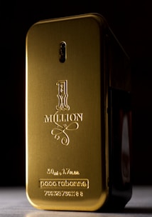 A gold-colored, rectangular perfume bottle with rounded edges and embossed text on its surface. The text includes '1 Million' along with the brand name and volume details. The bottle is set against a dark background, highlighting its shiny metallic finish.