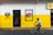 A person rides a bicycle past a building with a striking yellow and white facade. The building features a logo and the word 'Imperial' on the wall, alongside an advertisement depicting a red box with the same logo. There is a door leading into what appears to be a small shop.