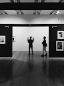 Two people stand in a gallery, observing black and white photographs on display. The setting is modern, with sleek wooden flooring and spotlights on the ceiling. The walls are labeled with city names and photographs are evenly spaced along them.