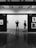 Two people stand in a gallery, observing black and white photographs on display. The setting is modern, with sleek wooden flooring and spotlights on the ceiling. The walls are labeled with city names and photographs are evenly spaced along them.