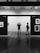 Two people stand in a gallery, observing black and white photographs on display. The setting is modern, with sleek wooden flooring and spotlights on the ceiling. The walls are labeled with city names and photographs are evenly spaced along them.