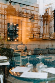 A sophisticated reflection photograph combining elements of interior and exterior views. The image features an ornate metal fence and architectural columns with warm light shining through, complimented by a plush, upholstered chair and a set of glassware on a table inside a cozy room. Artwork frames and illuminated bulbs add to the ambiance, blending exterior architectural details with an inviting interior seating arrangement.