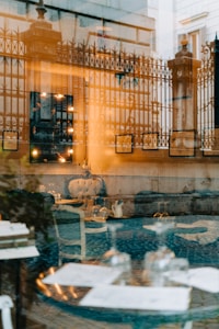 A sophisticated reflection photograph combining elements of interior and exterior views. The image features an ornate metal fence and architectural columns with warm light shining through, complimented by a plush, upholstered chair and a set of glassware on a table inside a cozy room. Artwork frames and illuminated bulbs add to the ambiance, blending exterior architectural details with an inviting interior seating arrangement.