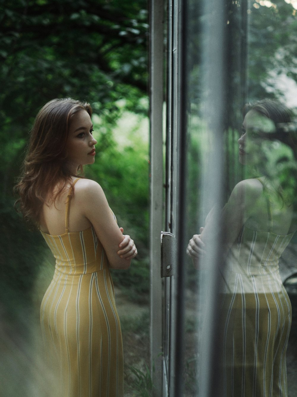 昼間にガラス窓の横に立つ黄色いスパゲッティストラップドレスの女性
