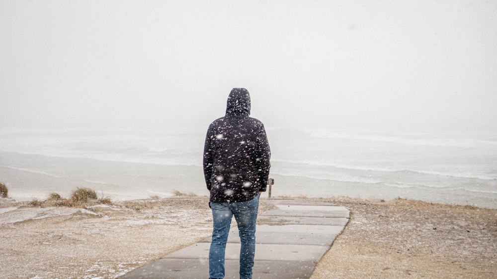 woman in black and white jacket and blue denim jeans standing on beach shore during daytime