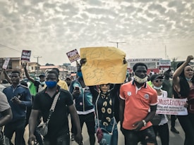 A group of people participating in a protest march, holding various signs and banners with messages such as '#END SARS BAD GOV'. Many participants are wearing face masks, and the mood appears to be serious and determined. The crowd is on a street with buildings in the background and a cloudy sky overhead.