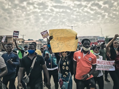 A group of people participating in a protest march, holding various signs and banners with messages such as '#END SARS BAD GOV'. Many participants are wearing face masks, and the mood appears to be serious and determined. The crowd is on a street with buildings in the background and a cloudy sky overhead.