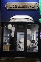 A barbershop storefront with a retro-style sign reading 'Dudes Barber Shop'. Inside, a person is working at a counter, wearing a face mask. The interior features a barber chair, shelves with various hair products, and a mirror on the right side.