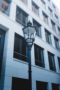 A black streetlamp stands in front of a modern building with a grid of large windows. The building has a simple, geometric design with white paneling. The lighting creates a cool and urban atmosphere.