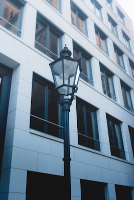 A black streetlamp stands in front of a modern building with a grid of large windows. The building has a simple, geometric design with white paneling. The lighting creates a cool and urban atmosphere.