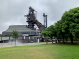 An industrial structure, possibly part of a historic steel mill, stands prominently with a mix of modern design elements. The area around it includes green trees and a well-maintained lawn. The sky appears overcast, contributing to a subdued atmosphere.