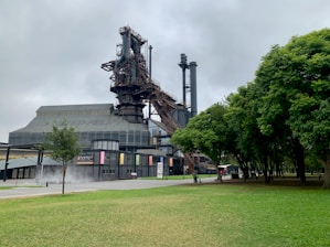 An industrial structure, possibly part of a historic steel mill, stands prominently with a mix of modern design elements. The area around it includes green trees and a well-maintained lawn. The sky appears overcast, contributing to a subdued atmosphere.
