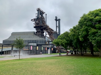 An industrial structure, possibly part of a historic steel mill, stands prominently with a mix of modern design elements. The area around it includes green trees and a well-maintained lawn. The sky appears overcast, contributing to a subdued atmosphere.