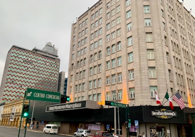 A city street with tall buildings, featuring a high-rise with numerous windows and a distinctive facade. Flags, including those of Canada, Mexico, and the United States, are visible near the entrance of a pub. A green traffic sign directs towards a shopping center. The atmosphere appears overcast.