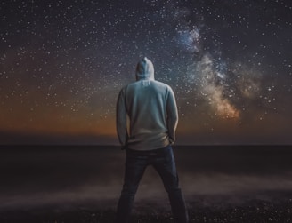 man in gray hoodie standing on black sand under starry night