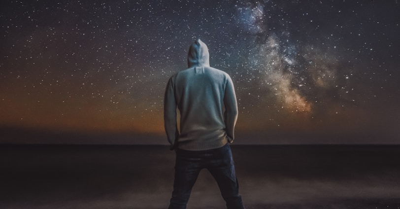 man in gray hoodie standing on black sand under starry night