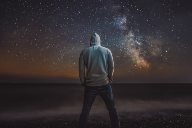 man in gray hoodie standing on black sand under starry night