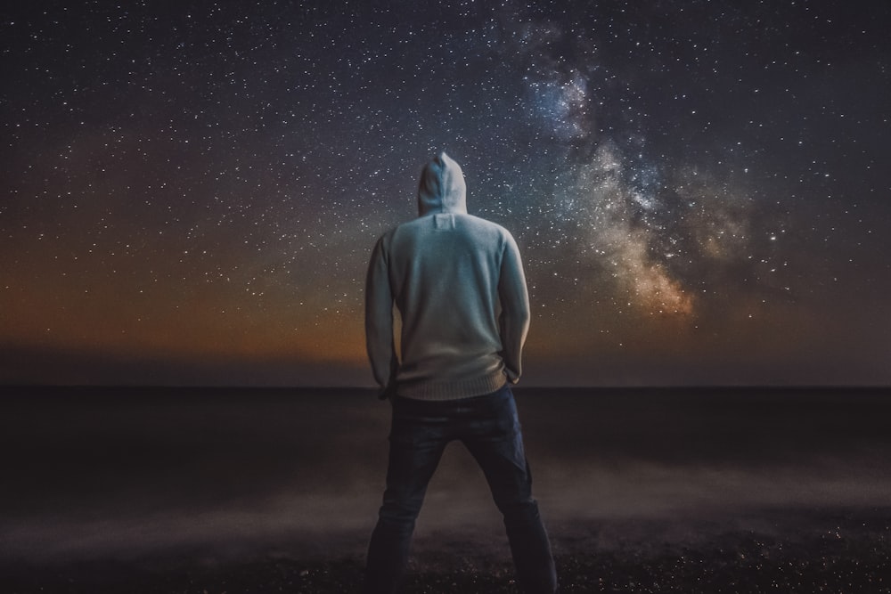 man in gray hoodie standing on black sand under starry night