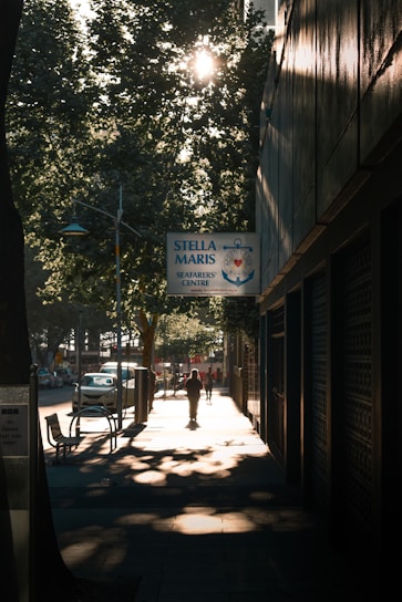 A sunlit street scene features a person walking along a sidewalk, with trees casting shadows. A sign for Stella Maris Seafarers' Centre is prominently displayed. Vehicles are parked along the street, and the sunlight creates a serene atmosphere.