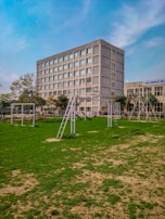 A multi-story rectangular building with numerous windows, surrounded by a grassy field with patches of dry soil. Several metal structures, resembling exercise or playground equipment, are positioned on the grass. Trees and bushes line the area near the building, which is set against a partly cloudy blue sky.