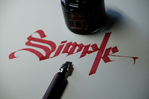 Red calligraphy spells the word 'Simple' on white paper. Next to the writing, there is a black ink jar and a calligraphy pen with a metallic nib.