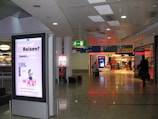 A public area inside what appears to be an airport or shopping mall features an illuminated advertisement with text and an illustration of a person using a laptop. The floor is shiny and reflective, and there are signs indicating directions to various gates or sections. A few individuals with luggage are walking through the corridor, and to the right, there are visible storefronts lit up, including a store with a sale sign.