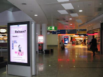 A public area inside what appears to be an airport or shopping mall features an illuminated advertisement with text and an illustration of a person using a laptop. The floor is shiny and reflective, and there are signs indicating directions to various gates or sections. A few individuals with luggage are walking through the corridor, and to the right, there are visible storefronts lit up, including a store with a sale sign.