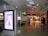 A public area inside what appears to be an airport or shopping mall features an illuminated advertisement with text and an illustration of a person using a laptop. The floor is shiny and reflective, and there are signs indicating directions to various gates or sections. A few individuals with luggage are walking through the corridor, and to the right, there are visible storefronts lit up, including a store with a sale sign.