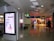A public area inside what appears to be an airport or shopping mall features an illuminated advertisement with text and an illustration of a person using a laptop. The floor is shiny and reflective, and there are signs indicating directions to various gates or sections. A few individuals with luggage are walking through the corridor, and to the right, there are visible storefronts lit up, including a store with a sale sign.