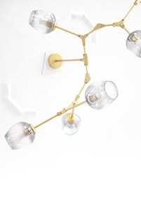 A modern, ceiling-mounted chandelier with a gold frame and multiple glass bulb covers. The design features articulated arms and clear glass coverings, creating an elegant and contemporary look against a white ceiling with decorative molding.