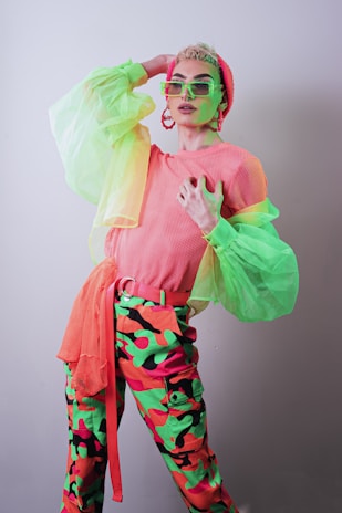 A person poses with one hand behind their head and the other on their chest, wearing bright, colorful clothing. The outfit includes a neon mesh top, a beanie, and trousers with a vivid camouflage pattern in green, pink, and orange. They accessorize with oversized glasses and earrings, and the clothing has striking neon green sleeves.