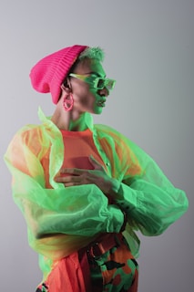 A person is wearing a bright and colorful outfit consisting of a neon green sheer top over a mesh fabric, paired with bold, patterned pants. Accessories include a vibrant pink beanie, oversized green glasses, and matching pink hoop earrings. The individual strikes a confident pose with one hand on the chest, set against a neutral background.