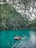 A person relaxes on an inflatable tube floating on a tranquil, turquoise lake surrounded by lush, green foliage. Overhanging branches create a natural canopy above the water.