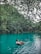 A person relaxes on an inflatable tube floating on a tranquil, turquoise lake surrounded by lush, green foliage. Overhanging branches create a natural canopy above the water.