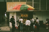 A small street food stall is covered by a multicolored umbrella. Several people are gathered around the stall, some standing and others sitting at a small table nearby. The setting appears to be in an urban area, with a partially visible sign and numbers on a building in the background.