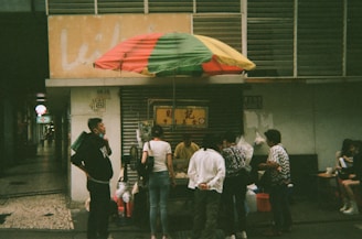 A small street food stall is covered by a multicolored umbrella. Several people are gathered around the stall, some standing and others sitting at a small table nearby. The setting appears to be in an urban area, with a partially visible sign and numbers on a building in the background.