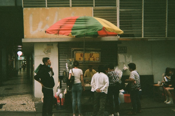 A small street food stall is covered by a multicolored umbrella. Several people are gathered around the stall, some standing and others sitting at a small table nearby. The setting appears to be in an urban area, with a partially visible sign and numbers on a building in the background.