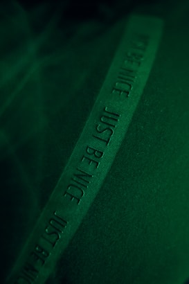 A dark green piece of fabric with an embossed text strip running diagonally across it, featuring the words 'JUST BE NICE'. The lighting creates a soft texture on the fabric surface.