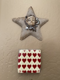 A wall-mounted star-shaped art piece features a textured net-like pattern with a ceramic face at its center. Below it is a square tile with a pattern of red hearts on a light background.