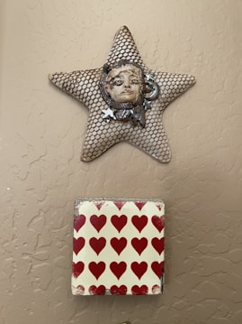 A wall-mounted star-shaped art piece features a textured net-like pattern with a ceramic face at its center. Below it is a square tile with a pattern of red hearts on a light background.