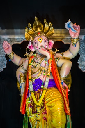 A beautifully adorned statue of an elephant-headed deity, decorated with vibrant colors and intricate jewelry, holding a variety of items in its multiple hands. The figure is draped in colorful garments and garlands, with a halo-like structure above its head, set against a dark background, emphasizing its divine presence.